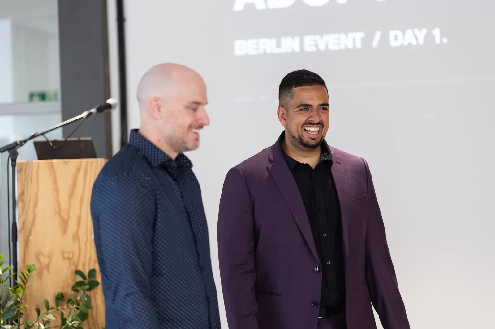 Mark Kashef laughing on stage at the Early AI-dopters Berlin event, Day 1 slide behind him